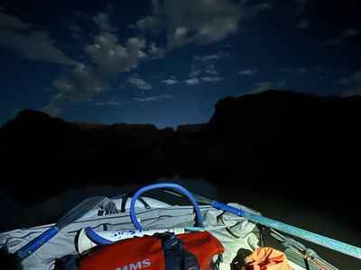 Night view forward over a loaded raft deck — oars, a red dry bag, a white bucket and a coiled blue hose in the foreground — toward dark canyon walls under a moonlit cloudy sky.