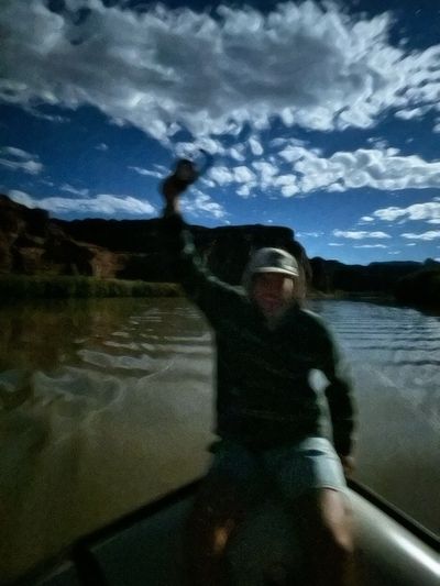 A heavily motion-blurred night frame of a crew member in a ball cap at the oars, one arm raised, with moonlit clouds above the dark canyon rim and the river behind.