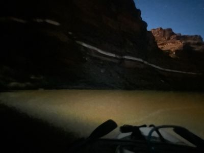 Night from the boat: a dark canyon wall with a pale seam of rock catching moonlight, a lit butte on the upper right under stars, and the raft's oar blade and frame silhouetted over ochre-lit water.