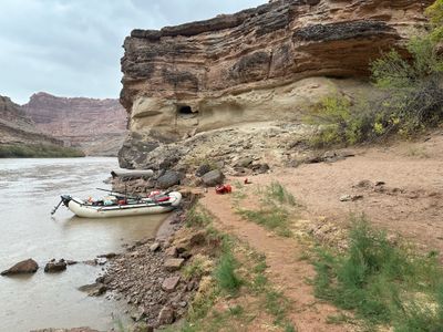A white raft with an oar frame, bimini top and small outboard motor sits beached on a muddy bank below an undercut sandstone wall with a small alcove, a red dry bag and orange gear on the red sand terrace under flat overcast light.
