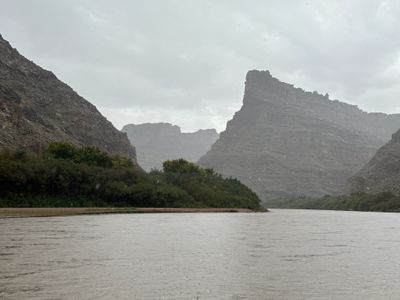 Raindrops speckle the lens over a wide flat gray-brown river running below layered talus slopes and a flat-topped caprock butte, with a low green brush-covered bar along the left bank under heavy overcast.