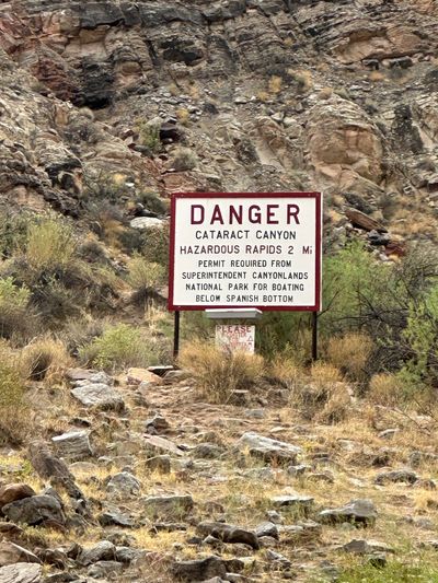 A white metal sign with a red border reading "DANGER — CATARACT CANYON — HAZARDOUS RAPIDS 2 Mi — PERMIT REQUIRED FROM SUPERINTENDENT CANYONLANDS NATIONAL PARK FOR BOATING BELOW SPANISH BOTTOM" stands on two posts on a dry, broken rock slope with a small weathered register box beneath it and dry grass and brush all around.
