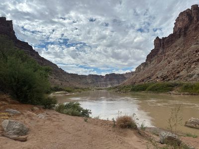 Wide view downstream from a red sand bank: the brown river curves left between a tall red sandstone buttress on the right and a rubble-and-cliff slope on the left, with layered gray and red canyon walls closing the view under broken cumulus.