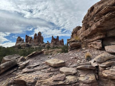 Clusters of banded red-and-white sandstone spires rise above a pinyon-juniper bench, seen across a foreground of stacked, broken slickrock ledges under blue sky streaked with high cirrus.