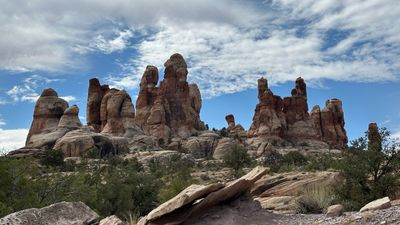 A wide stand of red-and-white banded sandstone spires and domes rises above a pinyon-juniper bench and pale slickrock, with a tilted slab in the foreground under blue sky and streaked cloud.