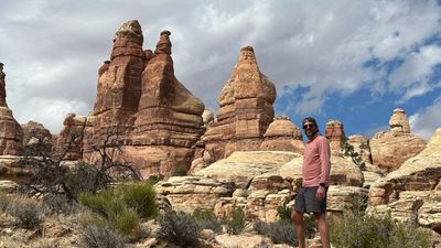 A bearded hiker in a pink long-sleeve sun shirt, cap and sunglasses stands on pale slickrock among towering banded sandstone spires, a dead juniper and sagebrush in the foreground under broken cloud.