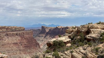 View from a juniper-dotted slickrock rim across a deep red-walled canyon to a hazy blue mountain range on the far horizon, with storm cloud spreading across the sky above it.