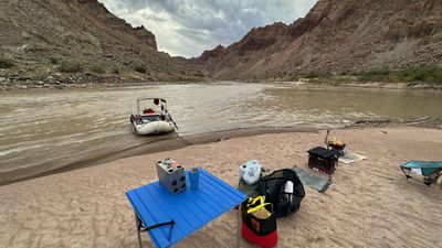 Camp on a sand bar at dusk: a blue roll-top table holding a camp stove and an insulated cup, dry bags and a black gear duffel on the sand, a fire burning in a metal fire pan on a ground cloth, a camp chair at right, and the bimini-framed raft tied off in the silty river below banded canyon walls.