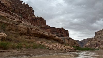 A long, tilted bench of red sandstone ledges rises from a muddy riverbank to a fluted cliff and canyon rim under solid gray overcast, with the silty tan river curving along the lower right of the frame.