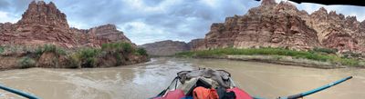 A stitched phone panorama shot from the rower's seat: the raft's gray bimini canopy and red and orange dry bags fill the bottom of the frame while the silty river runs downstream between green-fringed banks and pale red sandstone towers under a cloudy sky.