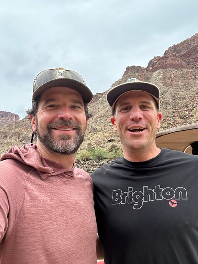 Two adult crew members in ball caps grin at a phone held at arm's length — one in a faded rose sun hoody with sunglasses pushed onto the brim, one in a black tee reading Brighton — with a gray talus slope, red canyon walls and the edge of the raft behind them.