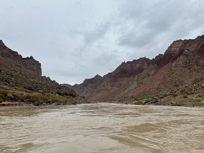 Looking downstream from water level at a wide, silty river running between steep gray-brown talus slopes and stepped red cliff bands, with boulder-piled banks on both sides under a flat overcast sky.