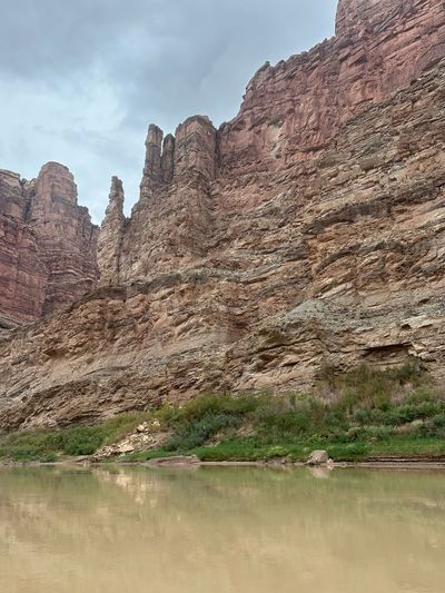 A slender detached sandstone pinnacle stands beside buttressed towers on a tall layered canyon wall, above a brushy green bank and a band of calm olive-tan river that mirrors the rock.