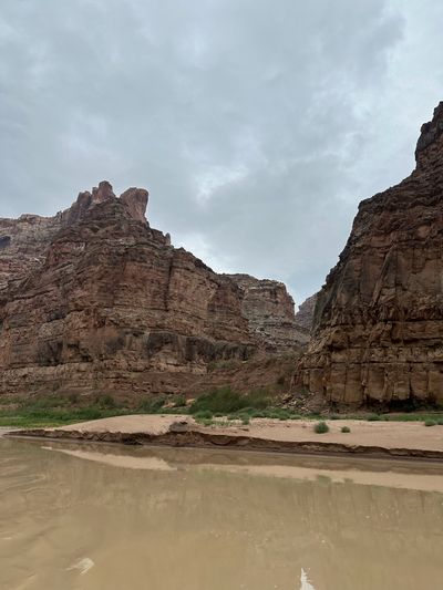 A wide sand bench with wind-rippled sand and a cut bank sits below stepped red canyon walls and a squared tower, with calm silt-colored water reflecting the cliffs across the foreground.