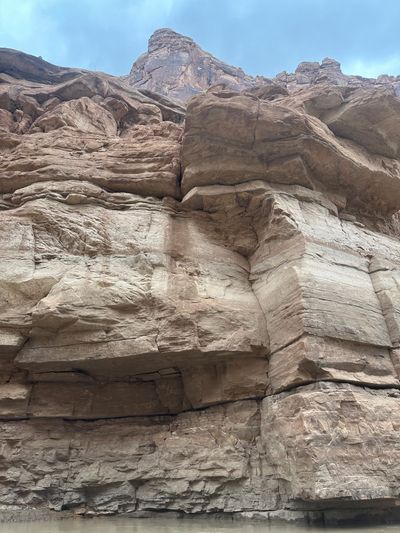 Looking steeply up a tall, blocky pale-tan sandstone wall rising straight out of the water, its ledges and vertical joints stacked below a dark red capping butte against a hazy blue-gray sky.