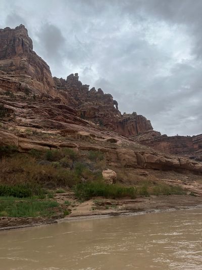 Weathered red sandstone benches with rounded potholes and knobby caprock climb toward a fluted cliff line under heavy gray cloud, with green riparian brush and a small sand fan meeting the muddy river along the bottom of the frame.