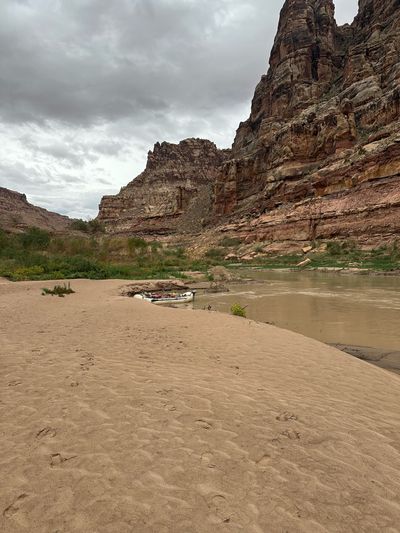 A rigged gray raft is nosed into the bank at the edge of a broad, footprinted sand beach, with the silty river curving past and tall red sandstone walls and a green side-canyon mouth rising beyond under heavy overcast.