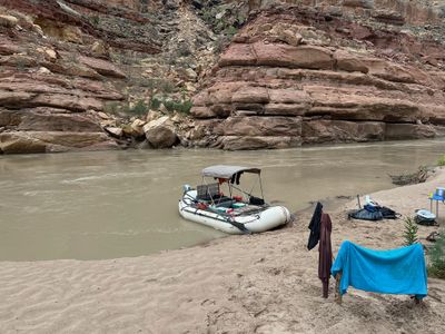 A rigged raft with a gray bimini canopy and a small outboard sits moored against a sand bar at camp, a blue towel and dark jackets hung to dry on driftwood posts, dry bags and life jackets set out on the sand and a blue camp table with pots at right, below ledgy red cliffs and a talus slope.