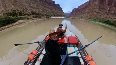 From a 360 camera mounted on the raft frame, the rower in a straw sun hat turns to the lens throwing hand signs while a second crew member on the bow raises a peace sign, blue oars trailing over glassy tan water between low canyon walls.