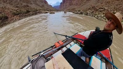 A fisheye frame from the boat: the rower in a straw hat and black top glances up at the lens from the oars, with orange dry boxes, coolers and rigging filling the deck and the silty river running out ahead between rocky banks.