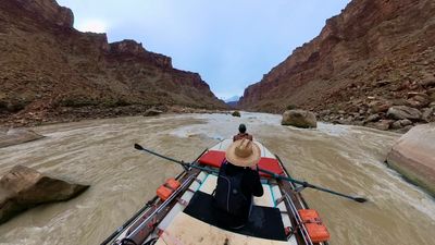 Seen from behind, the rower in a straw hat leans into the oars as the raft drops toward a boulder-choked rapid, whitewater breaking across the channel between talus banks and steep canyon walls.