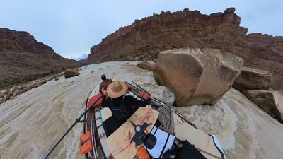 The raft slides past a house-sized tan boulder mid-rapid, brown water pillowing and breaking around the rock, with the rower in a straw hat working the oars and the canyon rim curving overhead in the fisheye frame.