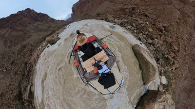 A near-overhead 360 frame looking straight down onto the raft as it runs a rapid, both crew, oars, dry boxes and rigging visible on the deck with churned brown-and-white water and rocky banks curling around the edges of the distorted frame.