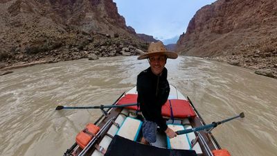 The rower stands at the oars in a straw hat and black drysuit top, grinning back at the boat-mounted camera, with the silty river opening ahead between rocky talus banks and red canyon walls.