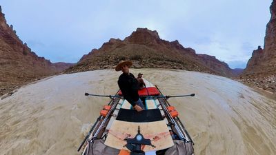 The rower in a straw hat looks back at the boat camera while standing at the oars, a pyramid-shaped red butte rising straight ahead over the churned tan river and rocky canyon slopes.
