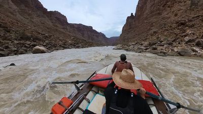 From behind the rower's straw hat, the raft enters a long white wave train between boulder banks, with a crew member sitting forward on the bow tube and the canyon corridor running out ahead.