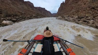 Deeper into the rapid, foaming brown-and-white water fills the channel ahead of the raft while the rower's straw hat and both extended oars frame the crew member crouched low on the bow.