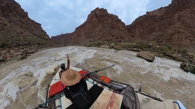 A crew member on the bow throws one arm up as the raft punches a breaking wave, spray flying off the left tube, with a broad red butte standing at the head of the canyon ahead.