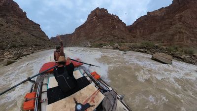 Water beads scatter across the lens as the raft drives through the wave, the bow crew member's arm still raised and whitewater curling along the left tube below the red butte.