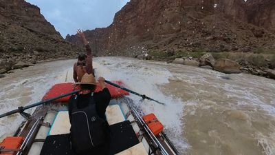 The bow crew member throws both arms overhead as the raft rides through a foaming wave, the rower braced on the oars in a straw hat and spray streaking across the lens.