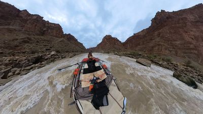 A wide, heavily distorted 360 frame of the raft running the rapid's white lane, the bow crew member's arm raised, gear strapped across the deck and the canyon walls bending around the edges of the shot.