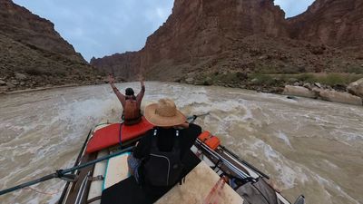 Seen from behind the rower's straw hat, the bow crew member holds both arms overhead as a wave breaks under the raft and washes down the right side of the boat.