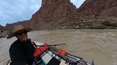 The rower leans toward the lens with a wide grin under a straw hat and dark sunglasses, one arm outstretched, the raft's red dry bag and orange boxes beside him and the silty river running along a talus slope beyond.
