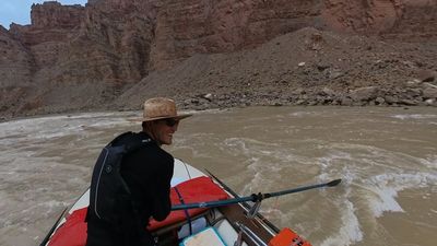 The rower in a straw hat and sunglasses smiles sideways at the boat camera, the raft's red dry bag and an oar filling the right of the frame and a gray talus slope below stepped red cliffs across the river.