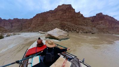 The raft lines up beside a large pale boulder parked mid-channel with water wrapping around it, the rower on the oars and a crew member forward on the bow, below a broad talus fan and a stepped red wall.