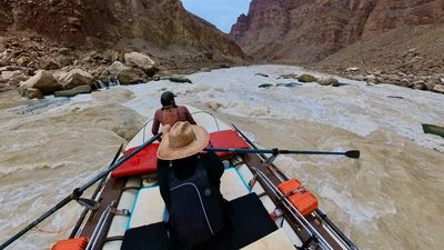 From behind the rower's straw hat, the raft drops into a long, bony rapid — dark rocks scattered through churning white water between boulder-piled banks under a gray sky.