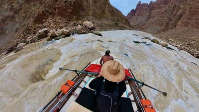 The raft runs a wide, boulder-strewn rapid with brown-and-white water breaking across the whole channel, the bow crew member low on the tube and distant spires standing on the skyline downstream.