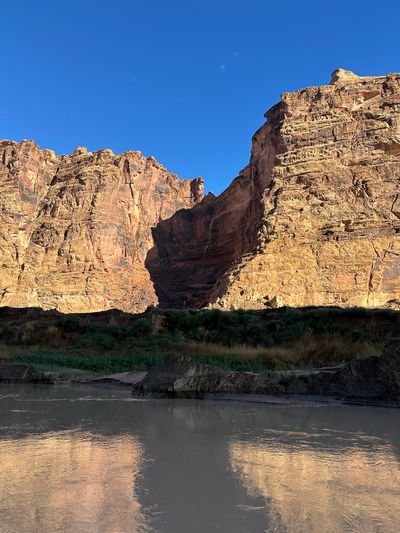 Two sunlit walls of layered tan and red sandstone meet at a shadowed side-canyon notch threaded by a thin ribbon of falling water, above a green brushy bench, dark mud banks, and calm silt-brown river reflecting the cliffs under a deep blue sky.