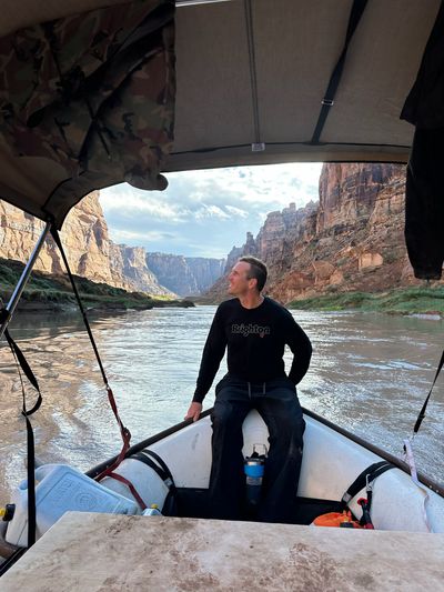 A crew member in a black long-sleeve shirt sits on the bow tube of a rigged raft under a tan bimini canopy, looking off to the right, with a white water jug, a blue insulated mug and an orange life jacket on the deck and the silty river running ahead between red canyon walls and towers.
