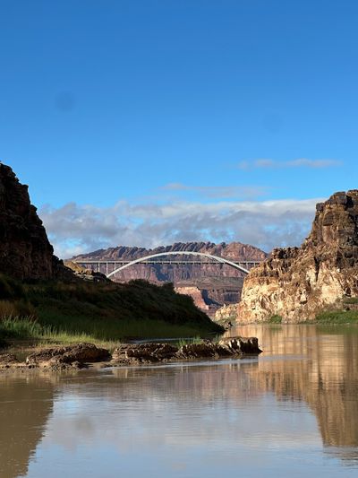A white steel arch highway bridge spans the canyon downstream, seen from river level across calm tan water, with mud banks and green reeds in the foreground and rounded red rock domes standing on the skyline under a blue sky.