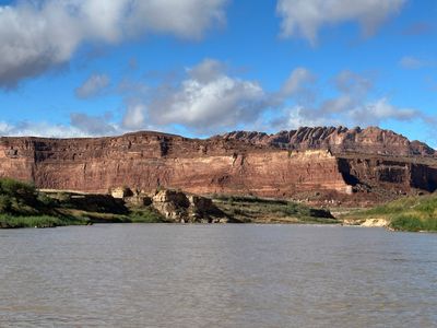 A long red cliff band topped by knobby rounded domes runs across the far bank above a green riparian bench and a low tan sandstone outcrop, with the wide silt-brown river filling the foreground under a blue sky and scattered cumulus.