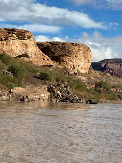A rounded tan sandstone bluff pitted with solution pockets and undercut by a shallow alcove rises above a sandy bank and a dark rock shelf fringed with brush, with distant red cliffs at right and the silty river running across the foreground.