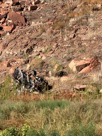A crushed and rusted car body lies wrecked on a red talus slope of broken rock and dry brush, with a thin cable strung across the hillside above it and green tamarisk filling the foreground.
