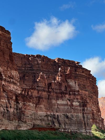 A tall red cliff face with a dark brown ledge band below the rim, where several protruding sandstone blocks are streaked bright white, standing above pale banded rock and a green brushy bank under a blue sky with cumulus.