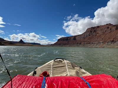 Looking over the tan-covered load and red gear rolls on the bow of a raft across choppy green reservoir water toward a red mesa wall stained with a pale waterline band at right and a distant sandstone tower at left, under towering white cumulus.