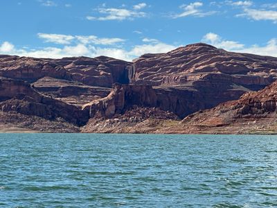 Stepped red sandstone cliffs and rounded slickrock domes rise across blue-green reservoir water, with a pale bleached waterline band and rubble slopes along the shoreline under a blue sky streaked with thin cloud.